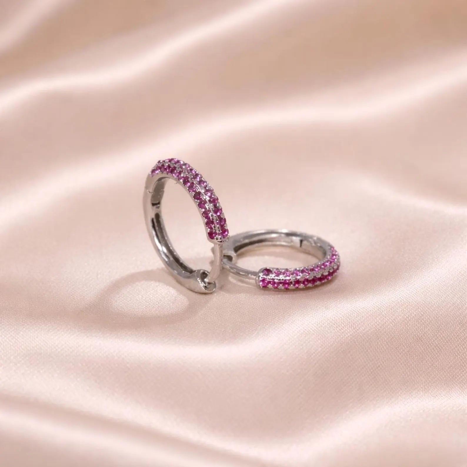 Pair of silver hoop earrings with pink gemstones on a pink satin background